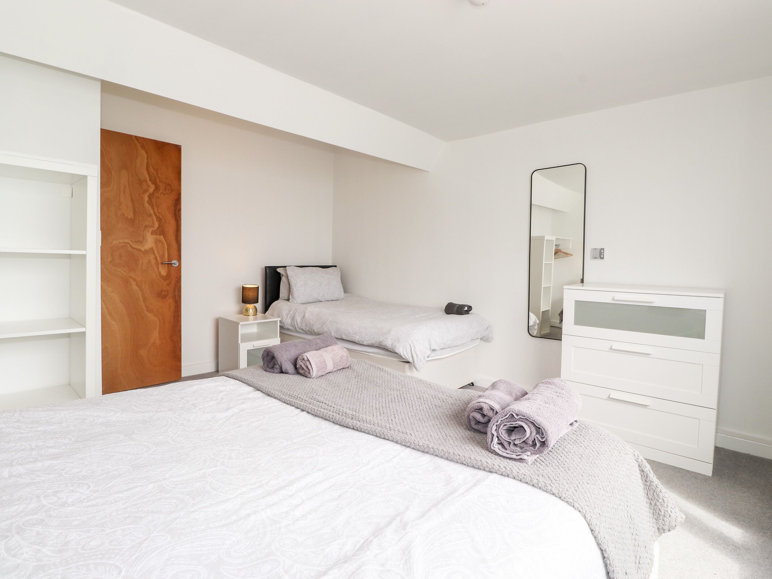 Un dormitorio con dos camas, una puerta de madera, una mesita de noche con lámpara, un espejo y un aparador blanco.