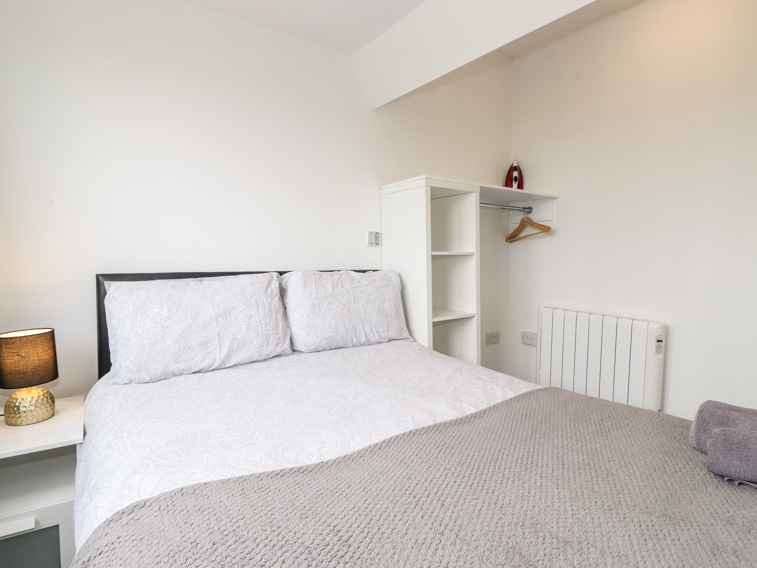 A bedroom with a bed, a bedside table with a lamp, a radiator, and a wardrobe with a hanger.
