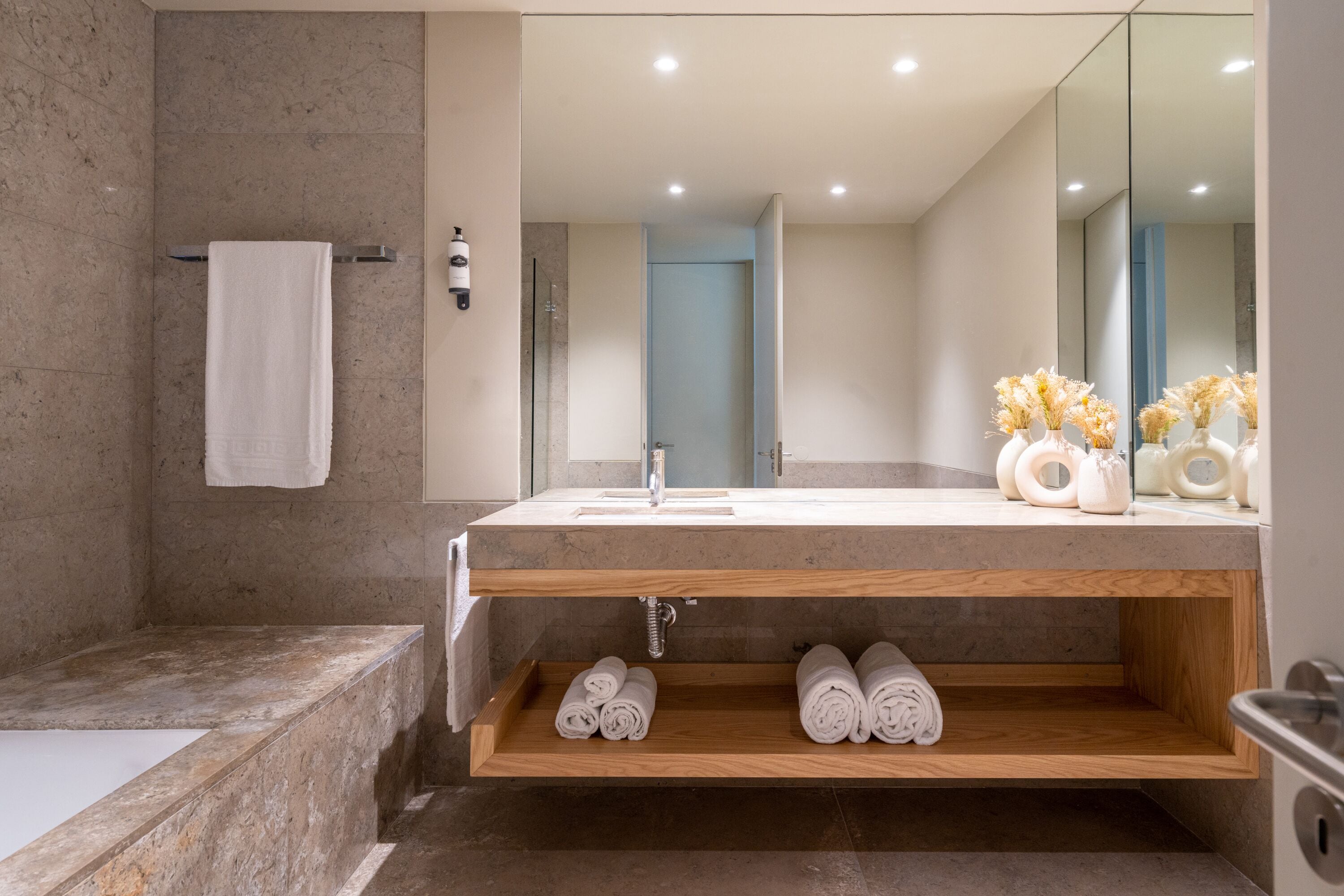 Une salle de bain moderne avec un lavabo en marbre, une étagère en bois où sont posées des serviettes et un grand miroir.