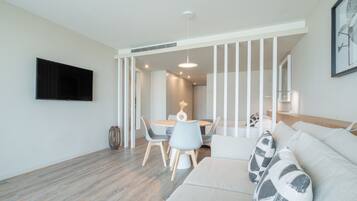 A modern living room with a flat-screen TV, a round dining table with chairs, a sofa with patterned cushions, and a staircase leading to another room.
