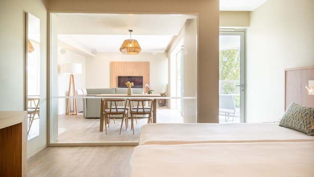 Une chambre d’hôtel moderne dotée d’un grand lit, d’un coin repas avec une table et des chaises, et d’un espace cuisine visible à travers une porte ouverte.