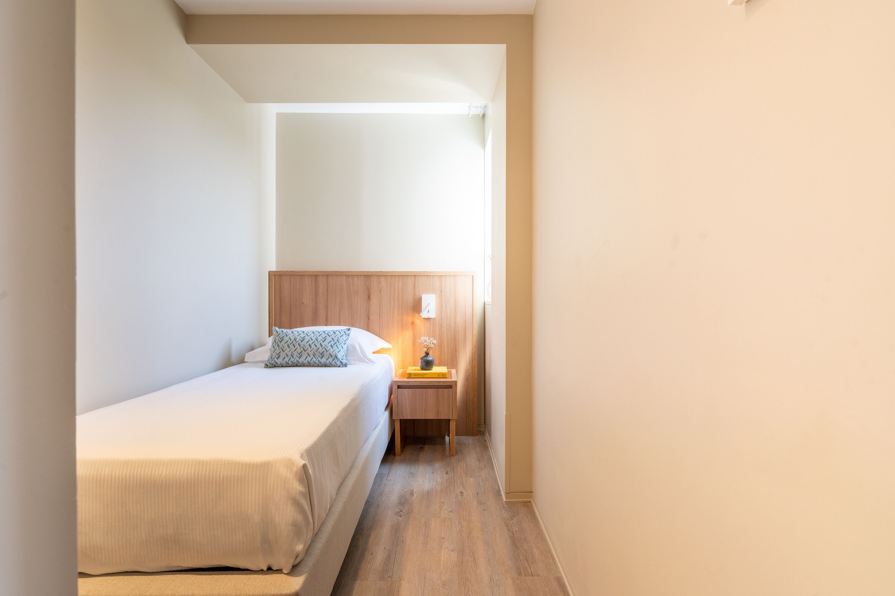 A narrow hotel room with a single bed, a nightstand with a lamp, and a wooden headboard.