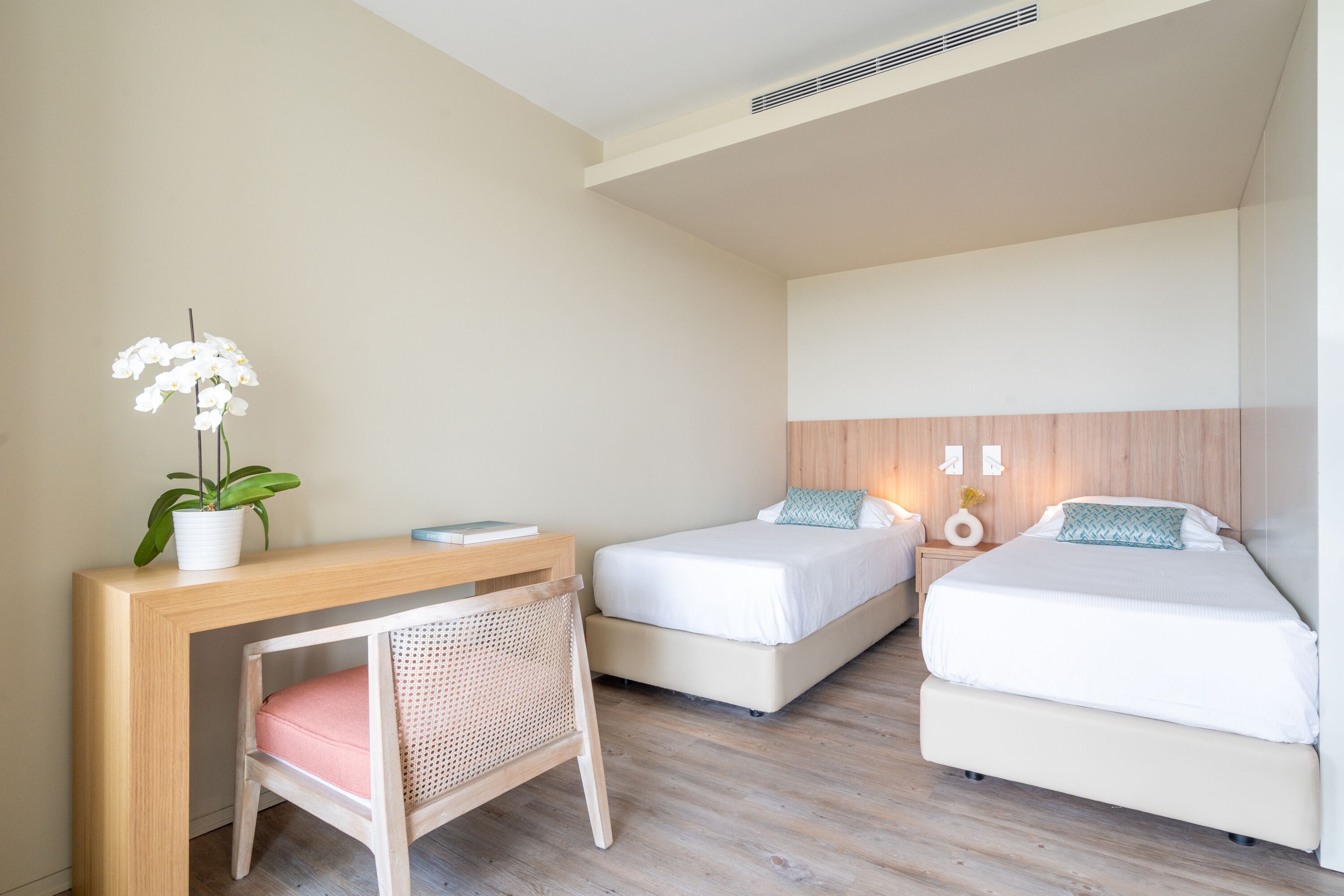 Quarto com duas camas de solteiro, uma escrivaninha com cadeira, um vaso de planta e uma mesa de cabeceira com abajur.