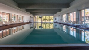 Une piscine intérieure moderne avec des parois en verre transparent, des chaises longues et une vue sur l’extérieur.