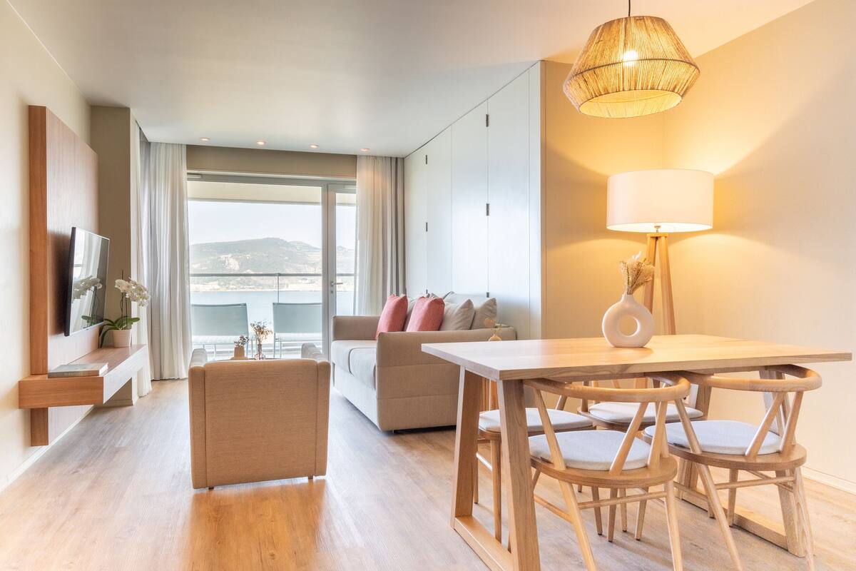 Ein modernes Wohnzimmer mit Sofa, einem Esstisch mit Stühlen, einem an der Wand befestigten Fernseher und einem großen Fenster mit Ausblick.