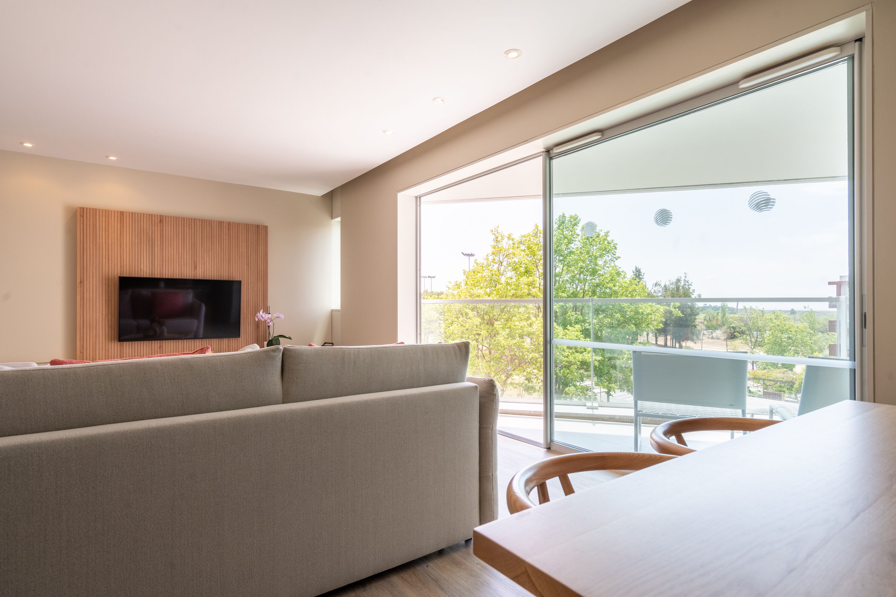 Uma sala de estar moderna com uma televisão de tela plana grande fixada em um painel de madeira, um sofá bege, uma mesa de jantar com cadeiras e uma grande porta de vidro deslizante que dá para uma varanda externa com vista para árvores e edifícios.
