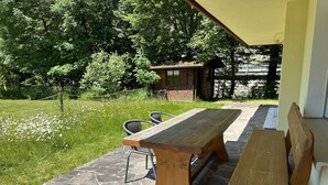 Un patio al aire libre con una mesa y bancos de madera, rodeado por un camino de piedra y abundante vegetación.