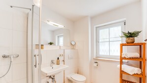 Un baño moderno con cabina de ducha de vidrio, un lavamanos blanco, un inodoro y un estante de madera con toallas.