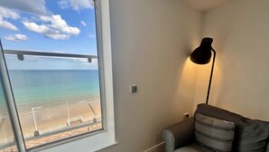 Et værelse med stort vindue med udsigt over en strand, en grå sofa med stribede puder, og en gulvlampe.