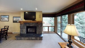 A living room with a stone fireplace, a flat-screen TV, and a large window with a view of trees. - Upscale 2-bedroom Residence in Resort-style Setting With Saline Outdoor Pool, 2 hot Tubs (Vail)