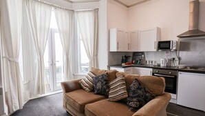 Ein kompakter Wohnraum mit Sofa, Küchenzeile und großen Fenstern mit Vorhängen.