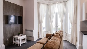 Ein modernes Wohnzimmer mit einer braunen Couch, einem flachbildfernseher an der Wand, einem kleinen weißen Tisch mit einer Pflanze und einem großen Fenster mit weißen Vorhängen.