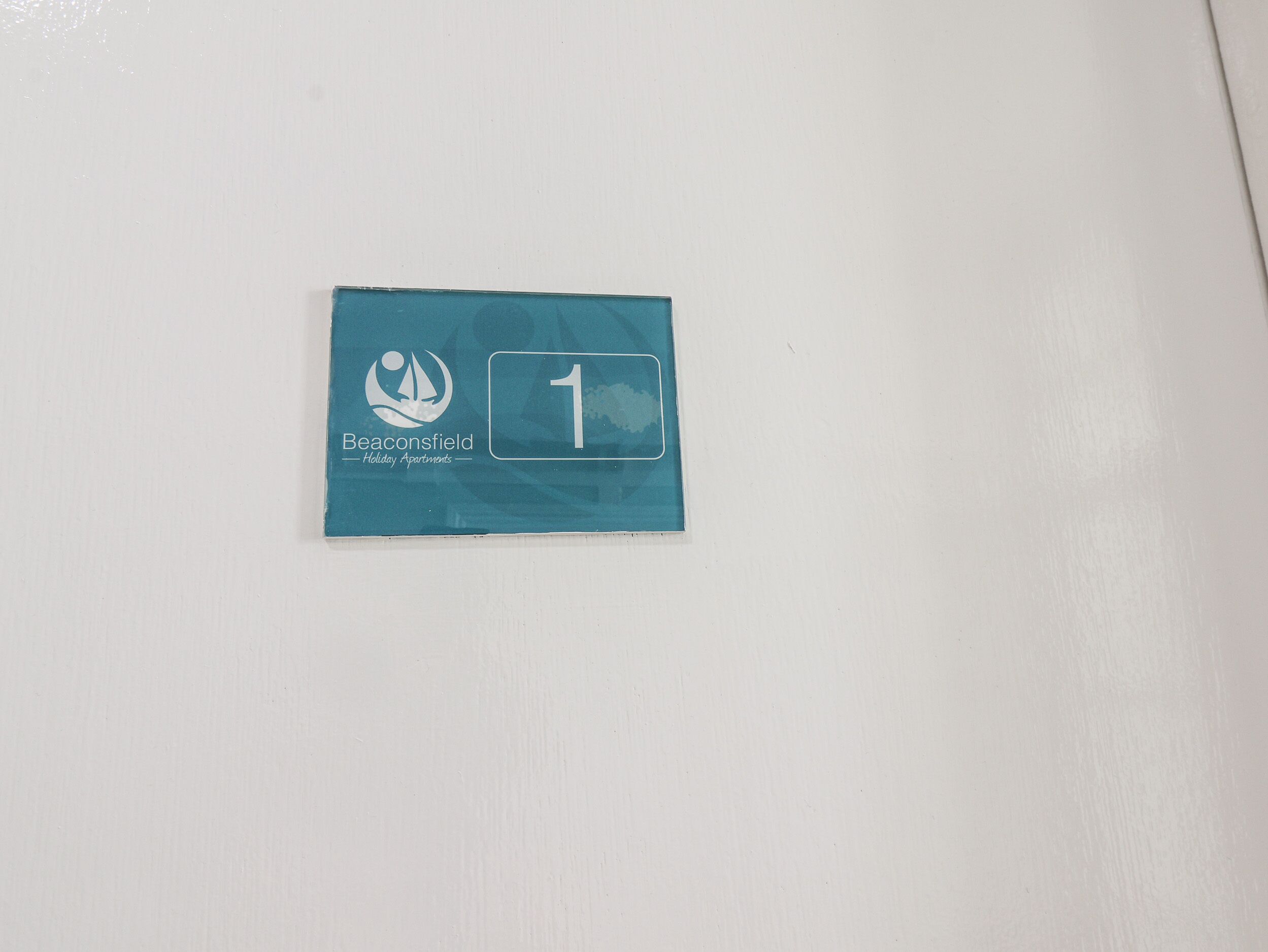 A small blue sign with the number 1 and the text "Beaconsfield Holiday Apartments" on a white wall.