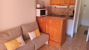 A compact kitchenette with a microwave, stove, and sink. A wooden cabinet and a small sofa with cushions are also visible. - Appartement Avec la Piscine Privée (Cavalaire-sur-Mer)