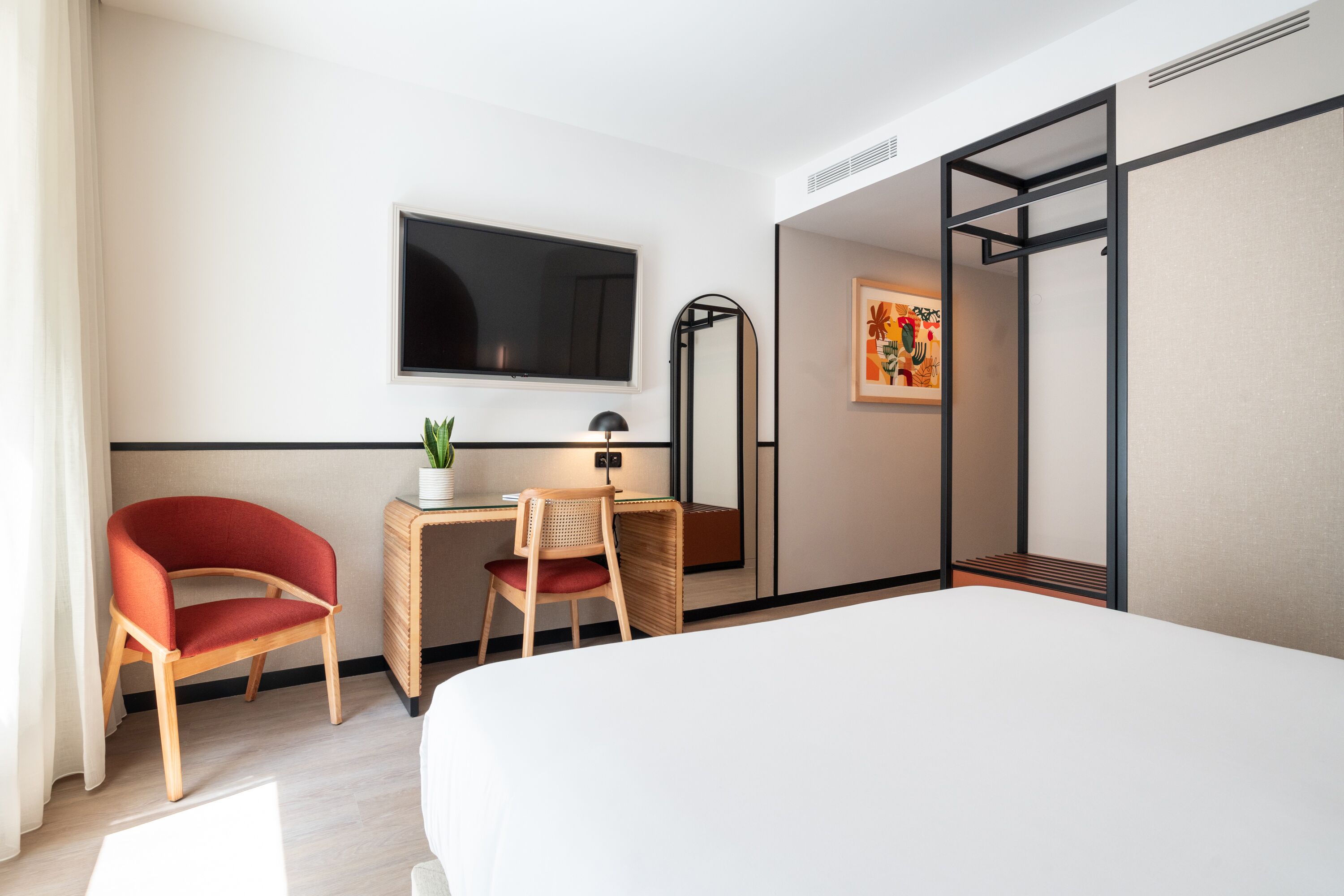 A hotel room with a bed, a desk with a chair, a TV, a mirror, and a painting on the wall.