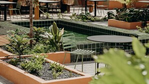 Un área de patio al aire libre con mesas y sillas de madera, una pequeña piscina con azulejos y diversas plantas.