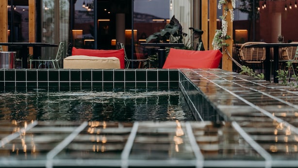 Un área de piscina con suelo de baldosas, rodeada de mobiliario exterior con cojines rojos y sillas de mimbre.