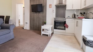 Un espace de vie compact comprenant un coin cuisine, un téléviseur à écran plat fixé à un mur en bois, une petite table blanche et un canapé gris.