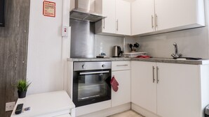 Une cuisine compacte avec des meubles blancs, une hotte de cuisine en acier inoxydable, un four encastré et un petit plan de travail sur lequel se trouvent une bouilloire et des ustensiles.