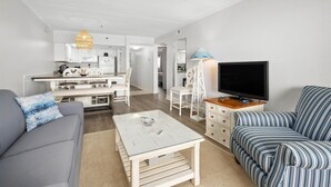 Un salon moderne avec un canapé, un divan à rayures, une table basse, un téléviseur et, au fond, un coin cuisine.