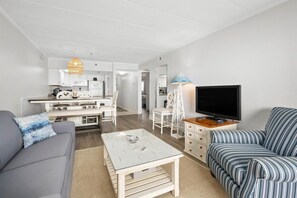 Un salon moderne avec un canapé, un divan à rayures, une table basse, un téléviseur et, au fond, un coin cuisine.