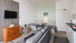 Quarto moderno com sofá, duas camas, televisão, cómoda de madeira, mesa pequena e área de cozinha.