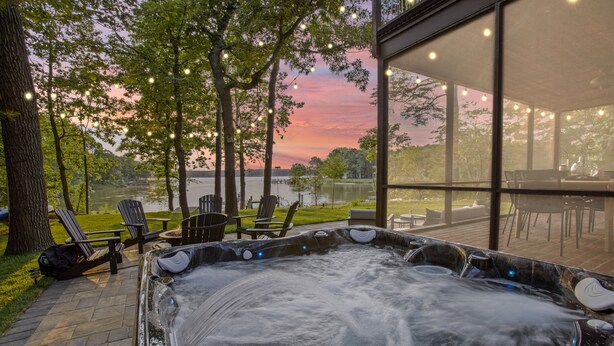 A hot tub with a view of a lake and sunset, surrounded by outdoor seating and trees. - Harbor View Haven | Waterfront w/ Hot Tub + Dock! (Chestertown)