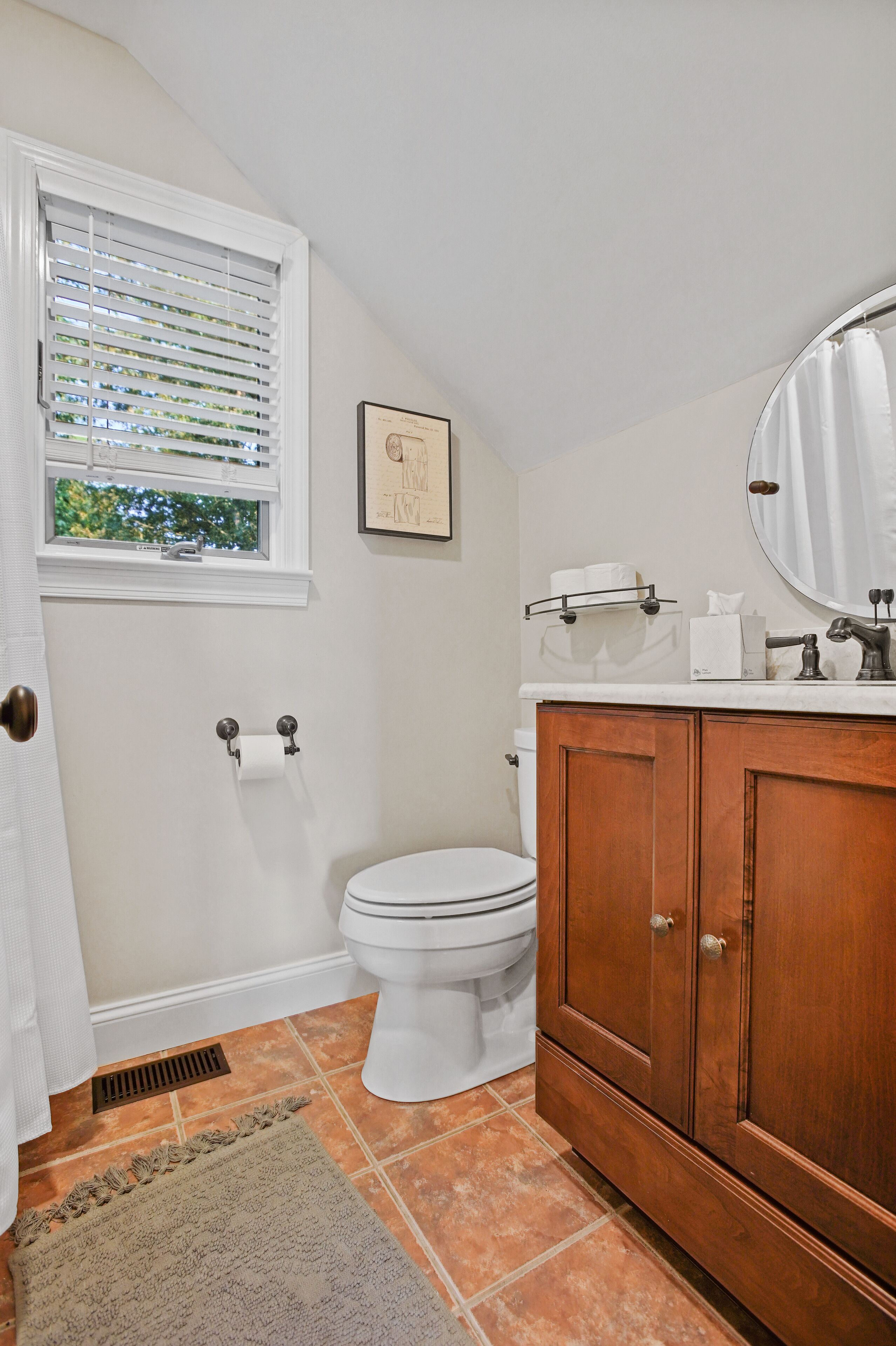A bathroom with a toilet, a wooden cabinet, a window with blinds, and a shower curtain.