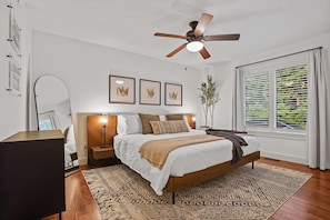 A bedroom with a bed, a ceiling fan, a nightstand, a bedside table, a lamp, a window with blinds, and framed artwork on the wall.