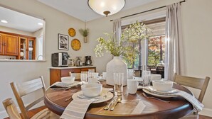 A dining area with a round table set for four, surrounded by wooden chairs. A vase with flowers is on the table, and there is a window with curtains behind it. - Harbor View Haven | Waterfront w/ Hot Tub + Dock! (Chestertown)