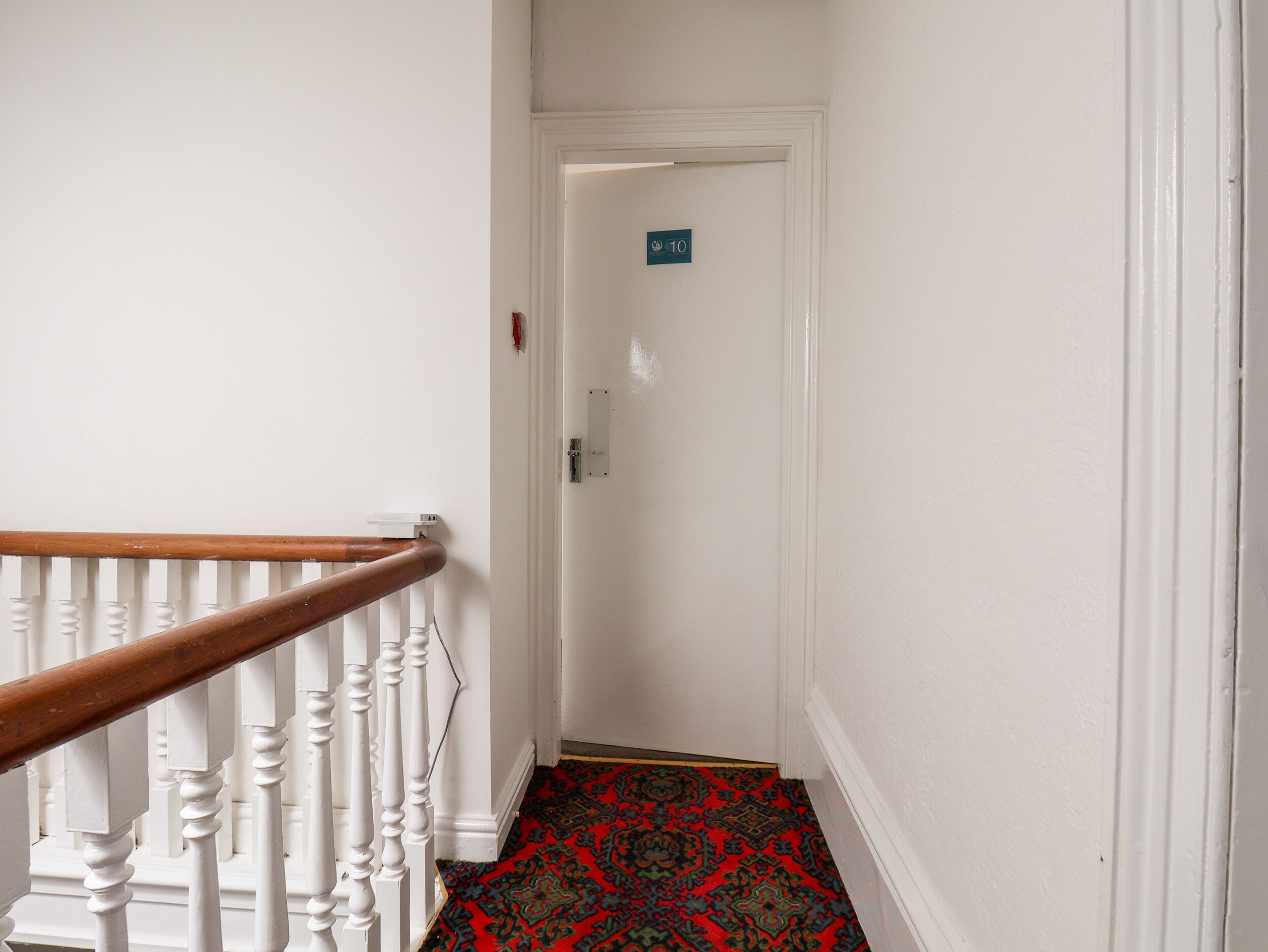Un couloir étroit aux murs blancs, avec une main courante en bois et un tapis à motifs. On aperçoit au loin une porte sur laquelle est apposée une pancarte.