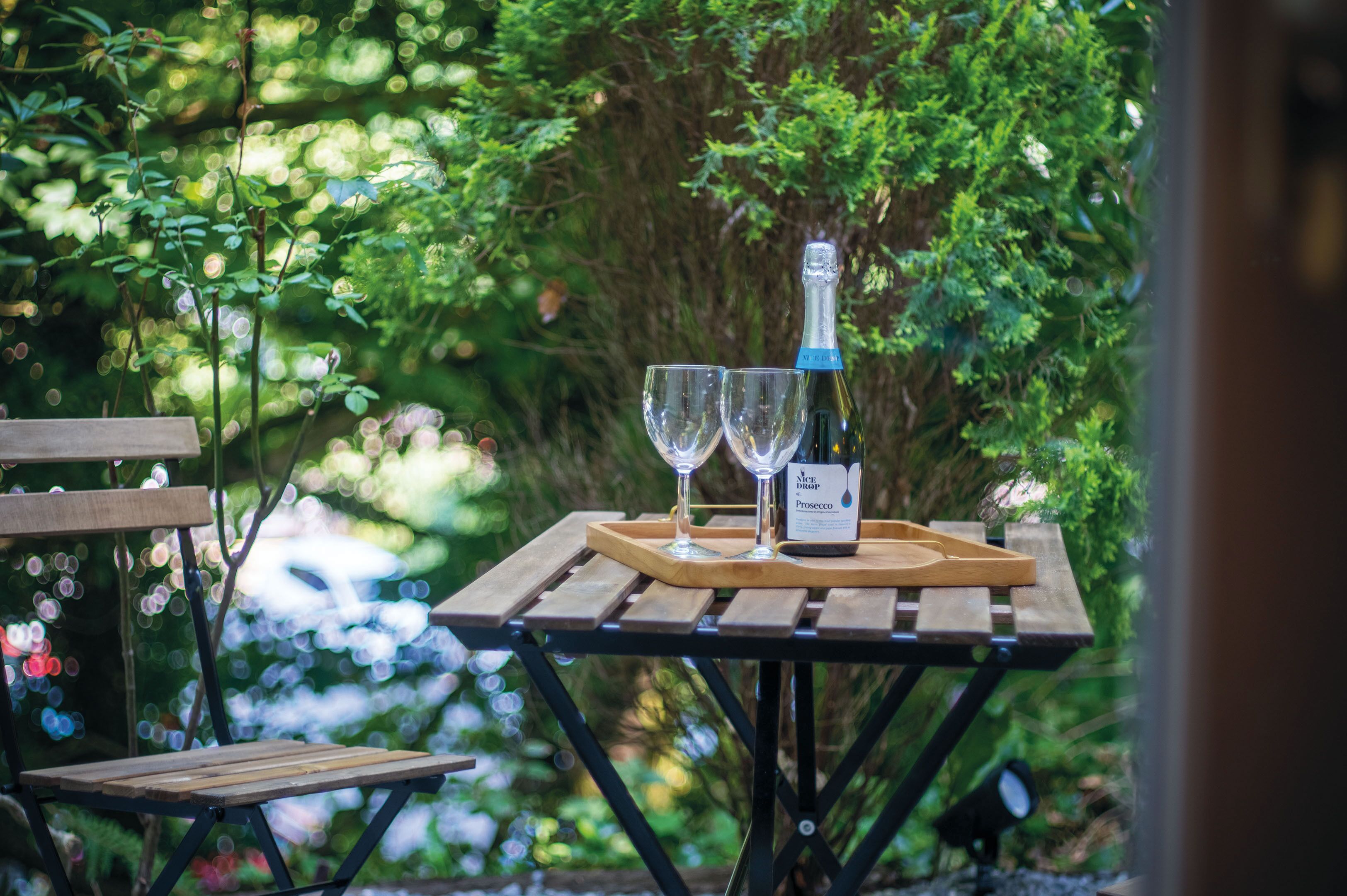 Une table en bois sur laquelle sont posés une bouteille de Prosecco et deux verres, le tout présenté sur un plateau, dans un décor extérieur agrémenté de verdure.