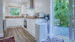 A modern kitchen with white cabinets, a built-in oven, a microwave, and a sink. There is a window with blinds, a door leading to a garden, and a small rug on the floor. - Croft Court - 1 Bedroom Apartment - Tenby (Tenby)