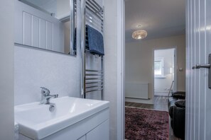 A bathroom with a white sink, a towel rack, and a mirror. There is a door leading to another room with a radiator and a patterned rug. - Croft Court - 1 Bedroom Apartment - Tenby (Tenby)