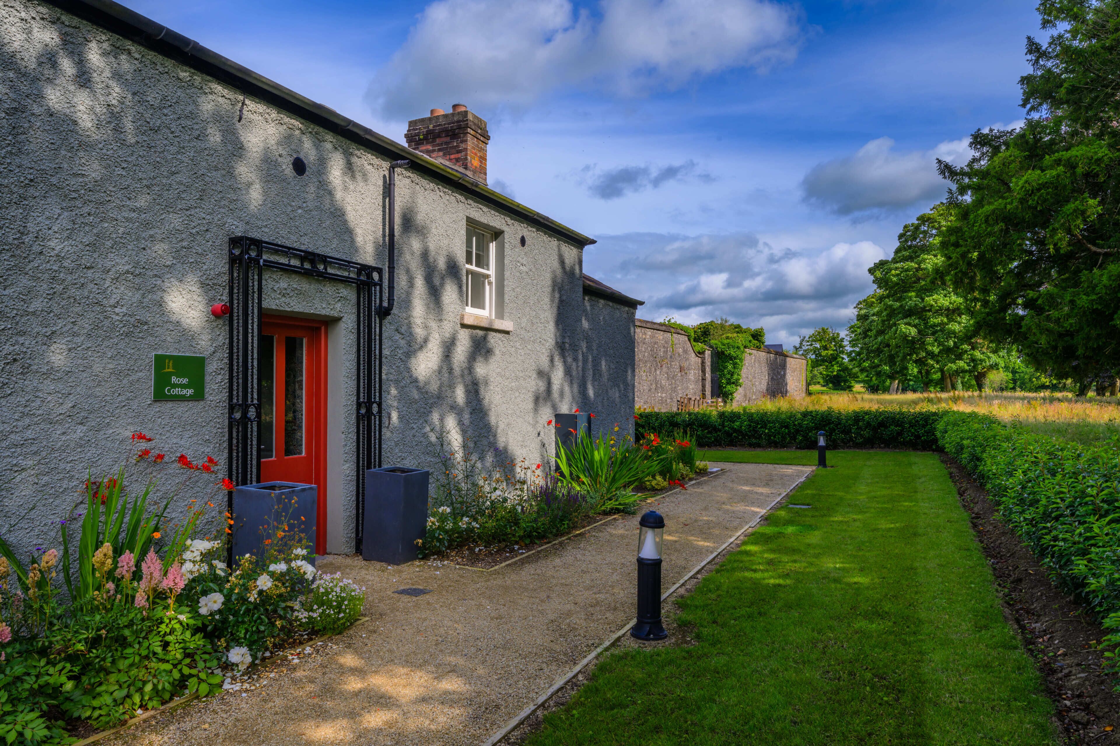 A stone cottage with a red door, a small window, and a gravel path leading to a grassy area.