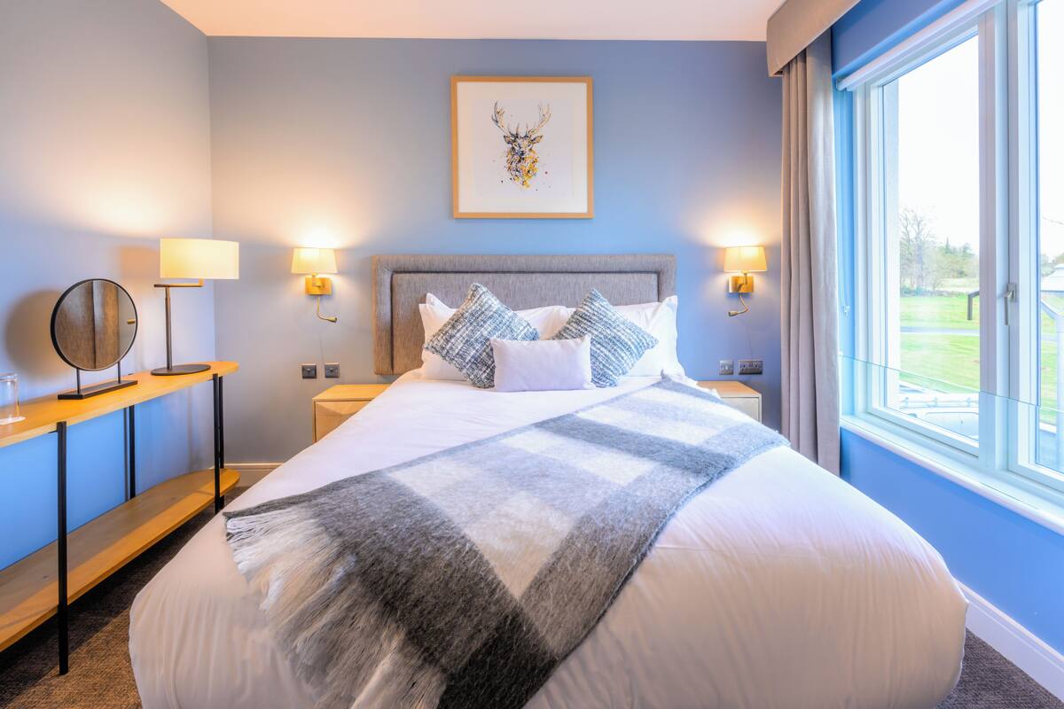 A neatly arranged bedroom with a large bed, two bedside lamps, a framed artwork above the bed, and a view of the outdoors through a window.