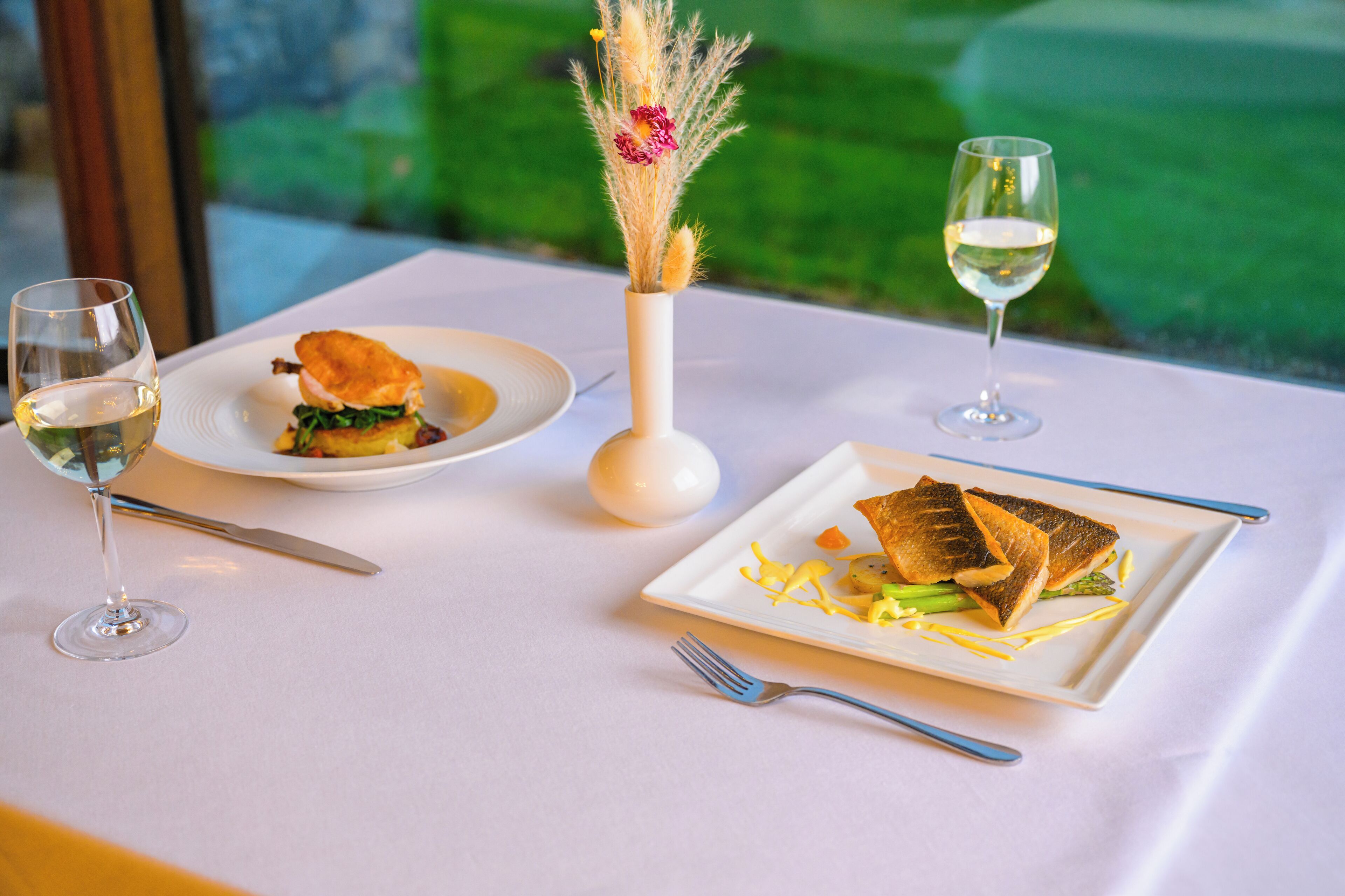 A table with a white tablecloth, two plates of food, a vase with dried flowers, and two glasses of white wine.
