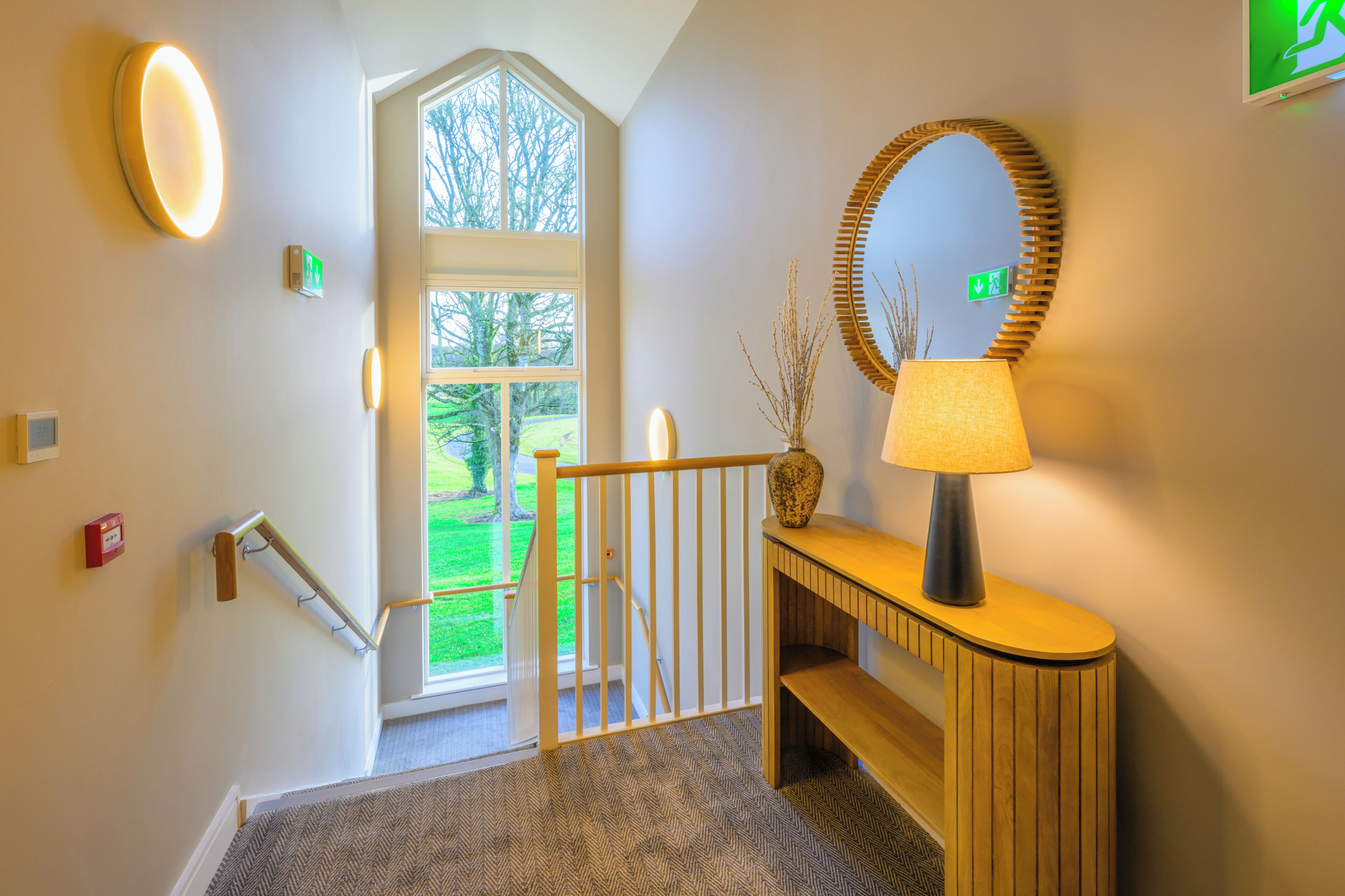 A staircase with a wooden handrail, a wall-mounted mirror, a lamp, and a door leading outside.
