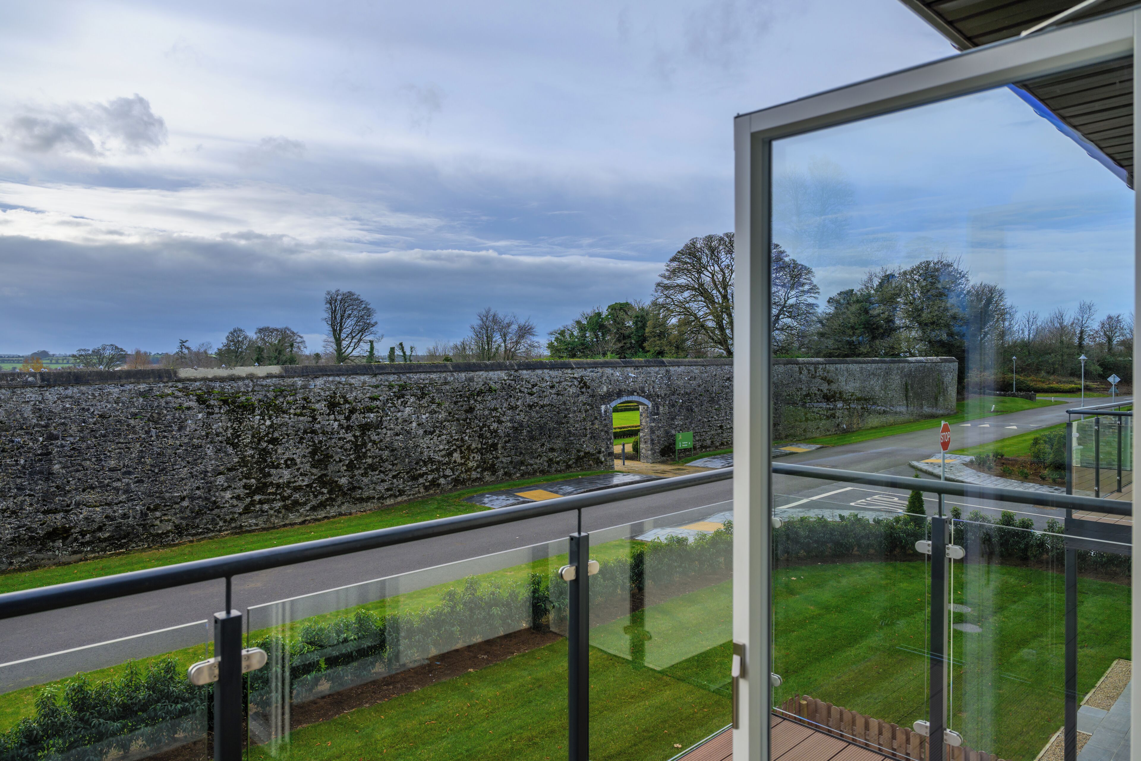 A view from a balcony with a stone wall, a grassy area, and a road with a stop sign.