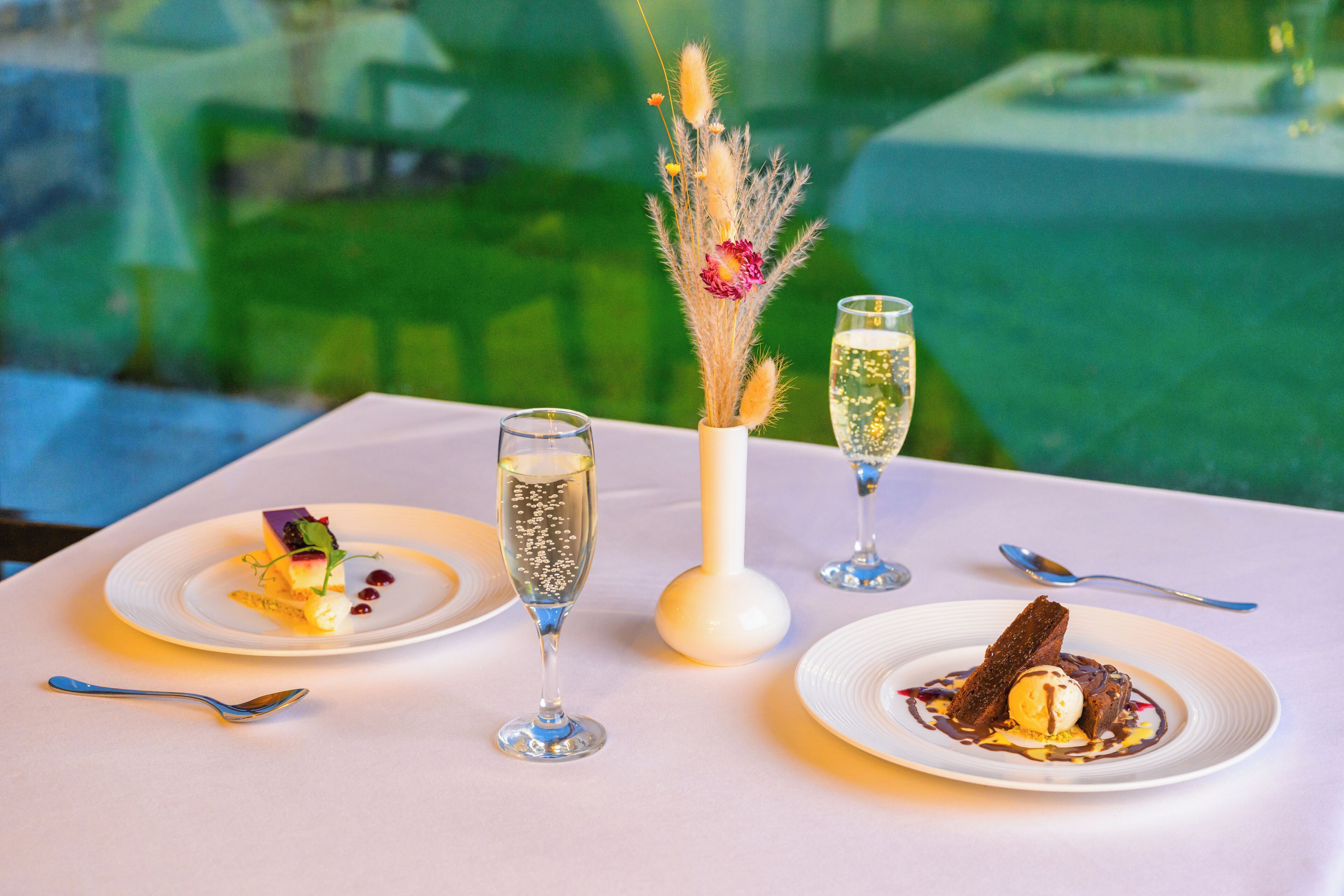 A table with three plates of dessert, two glasses of champagne, and a vase with dried flowers.