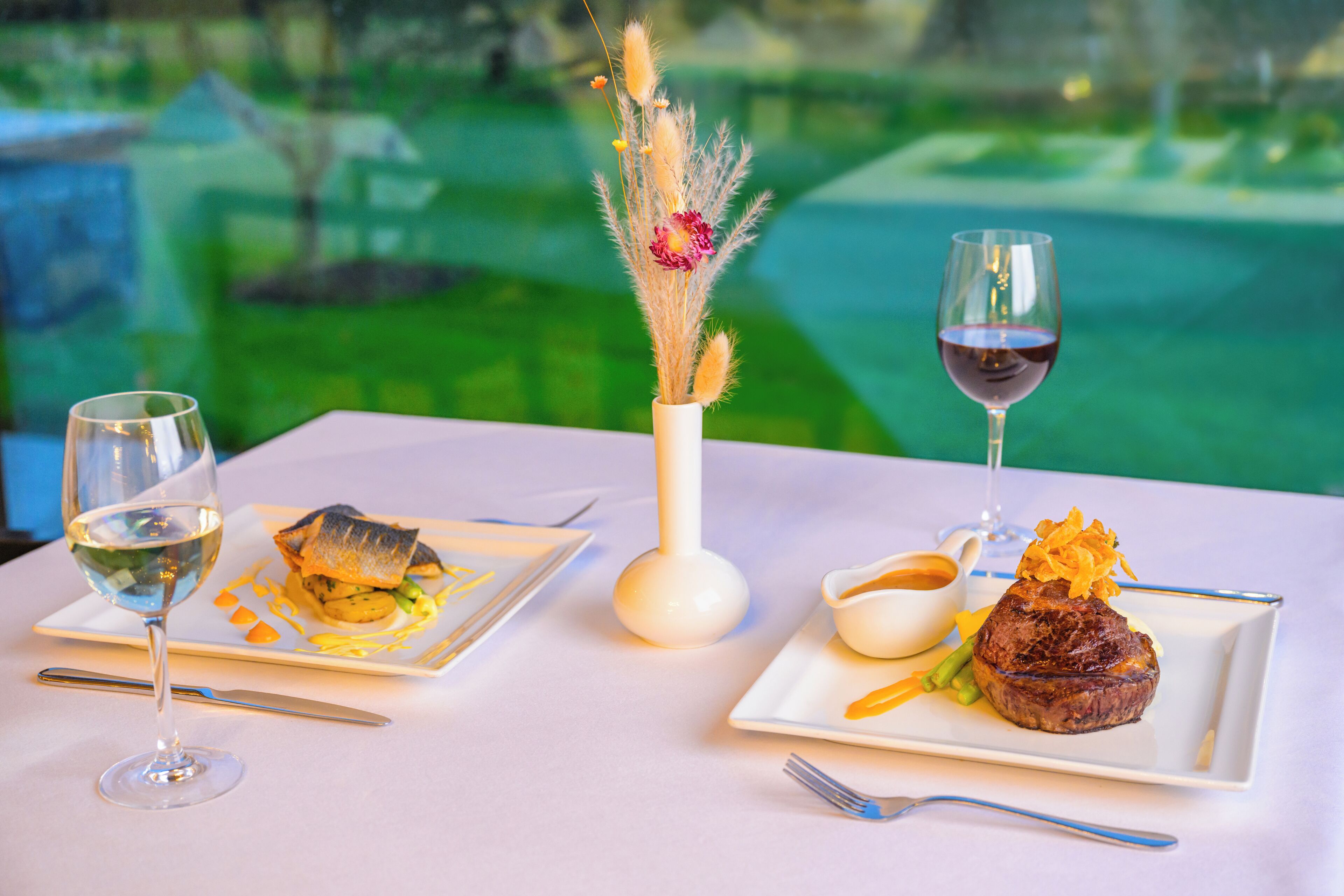 A table set with a white tablecloth, two square plates with food, a vase with dried flowers, and two glasses of wine.