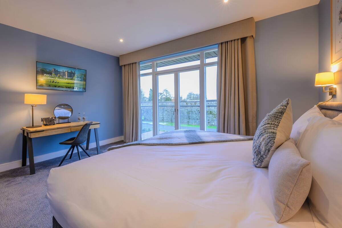 A hotel room with a large bed, a desk with a chair, a TV mounted on the wall, and a window with curtains.