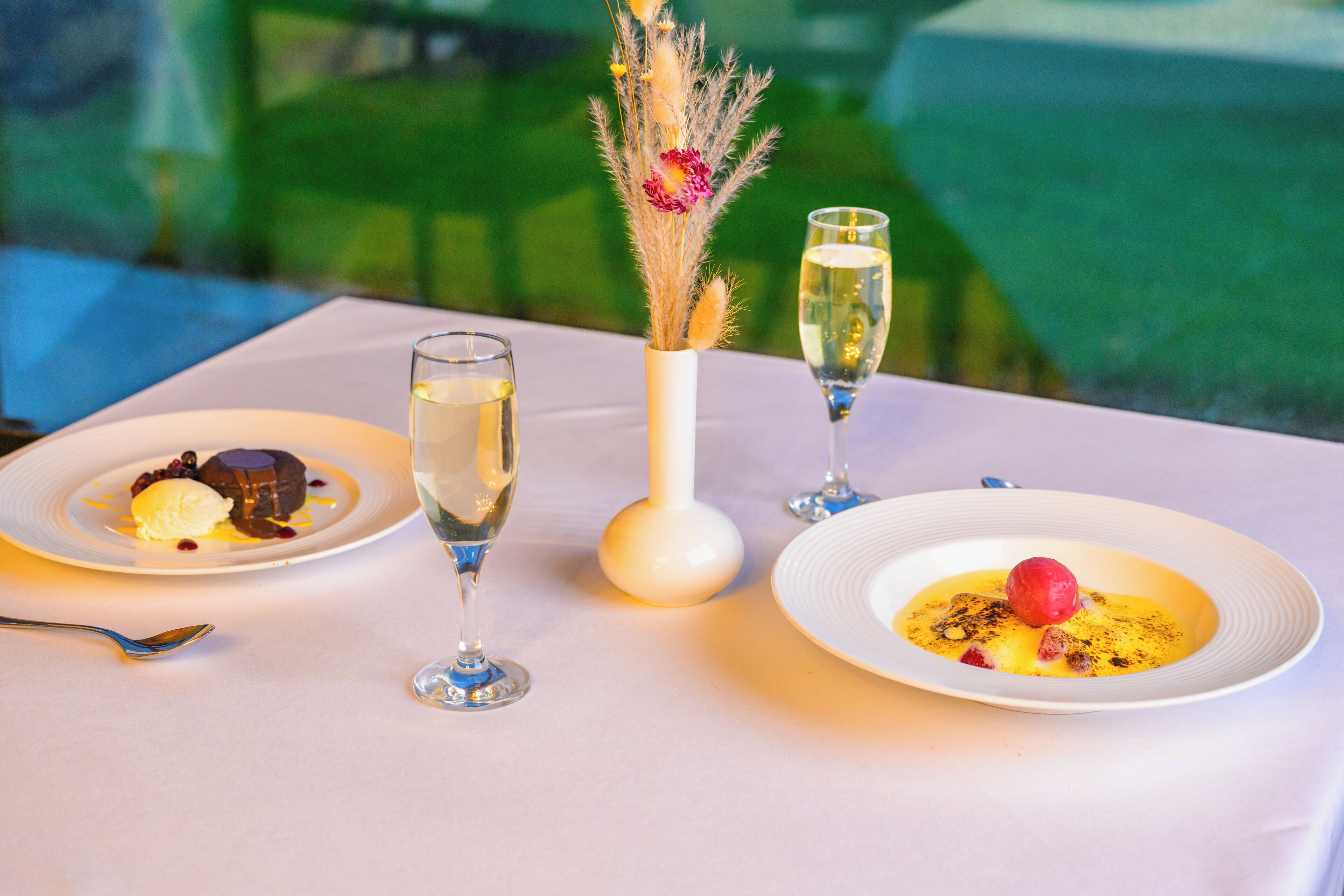 Un tavolo con due porzioni di dessert, due bicchieri di champagne e un vaso con fiori secchi.