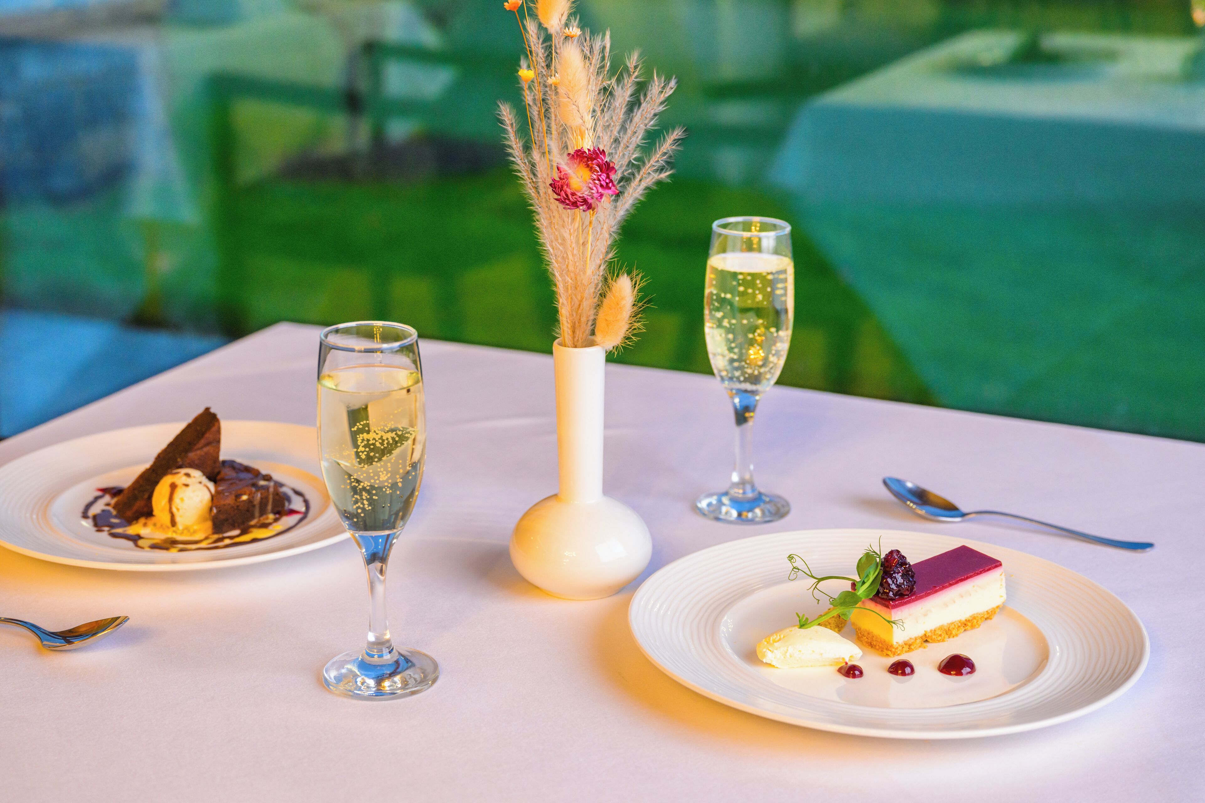 Una mesa con tres platos de postre, dos copas de champán y un florero con flores secas.