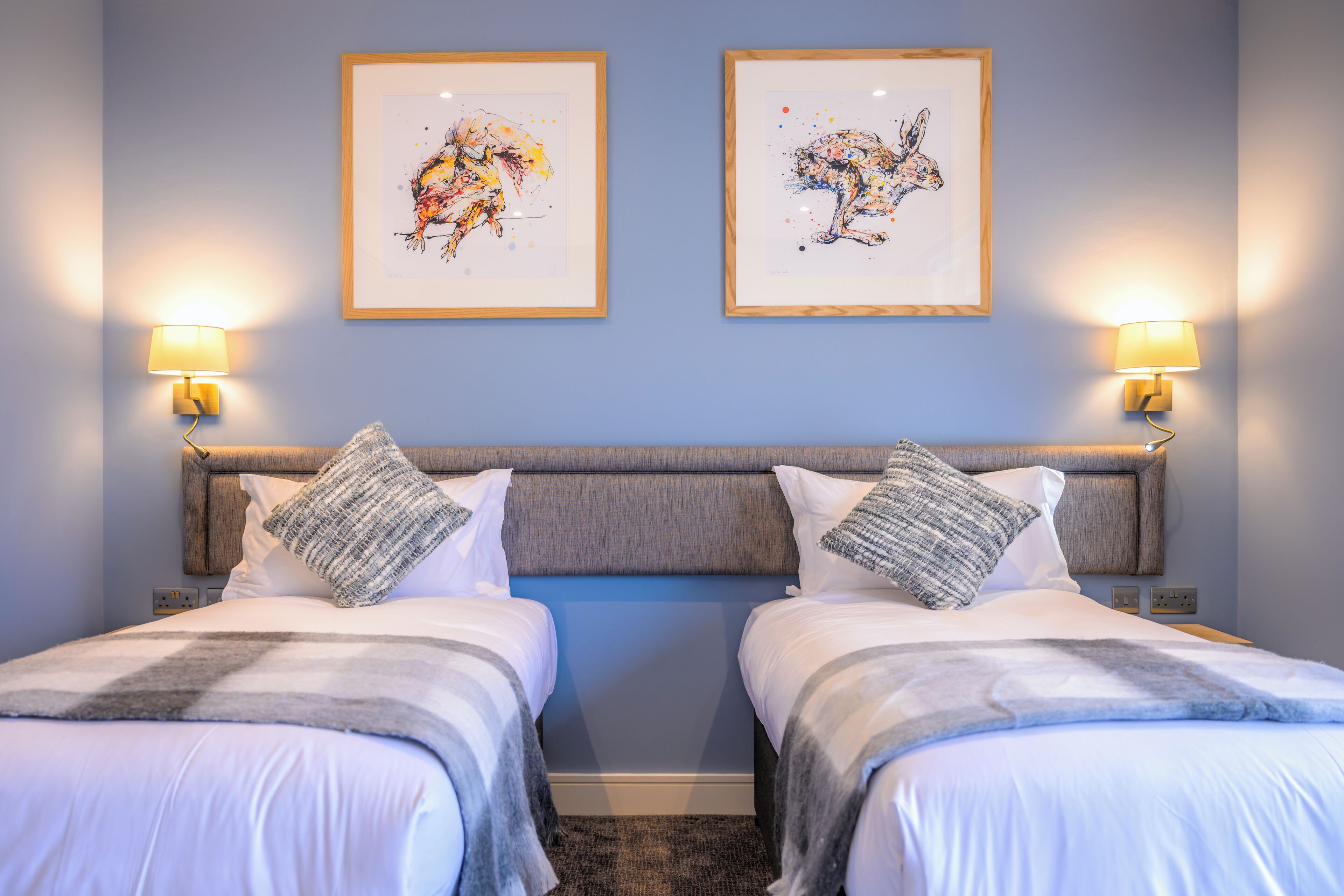 Habitación de hotel con dos camas, un cabecero gris y dos cuadros enmarcados en la pared.