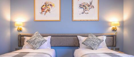 Habitación de hotel con dos camas, un cabecero gris y dos cuadros enmarcados en la pared.
