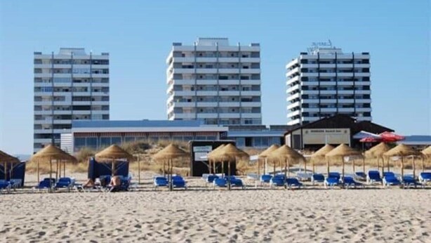Een strand met blauwe ligbedden en rieten parasols, een gebouw met een bord en hoge woongebouwen op de achtergrond.