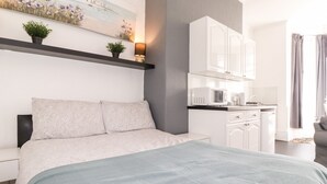 Une chambre moderne comprenant un lit, des tables de chevet, une commode, un bureau intégré, un petit coin cuisine et une fenêtre avec des rideaux.