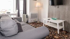 Un salon moderne avec un canapé gris, une table d’appoint blanche, un téléviseur à écran plat fixé sur un mur en bois, une lampe sur pied et un tapis à motifs.