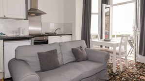 Un espace de vie compact comprenant un coin cuisine, un canapé gris avec des coussins, une table à manger avec des chaises et une vue sur l’extérieur grâce à une grande fenêtre.